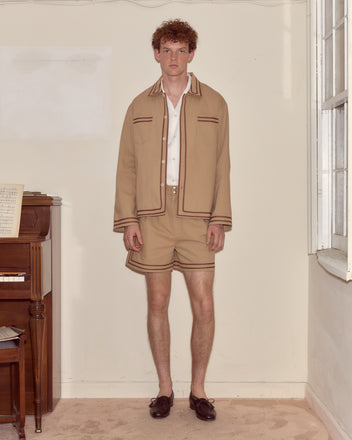 A man in beige shorts and a Canteen Overshirt stands indoors by a piano and window.