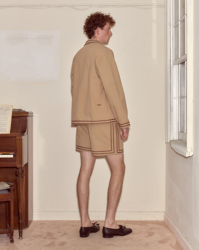 A man in tan shorts and the Canteen Overshirt, crafted from heavyweight cotton twill, stands indoors facing a corner by a window and piano.