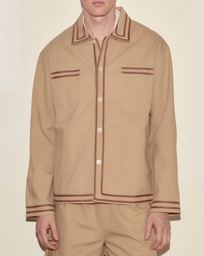 A person wears the Canteen Overshirt, a tan heavyweight cotton twill button-up with brown trim, paired with matching tan pants, standing against a plain background.