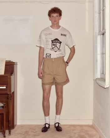 A man stands indoors by a piano, wearing a printed t-shirt, black Mary Jane shoes, and Canteen Shorts made from heavyweight cotton twill.