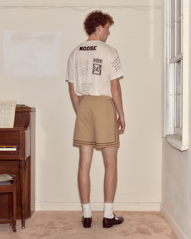 A man stands indoors, facing away, wearing a t-shirt, Canteen Shorts made of heavyweight cotton twill, white socks, and black shoes.