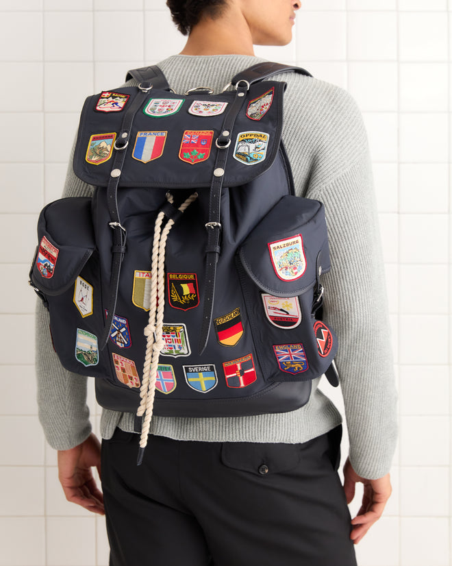 A person stands against a tiled wall wearing the Interrail Rucksack, featuring navy fabric and colorful embroidered patches.