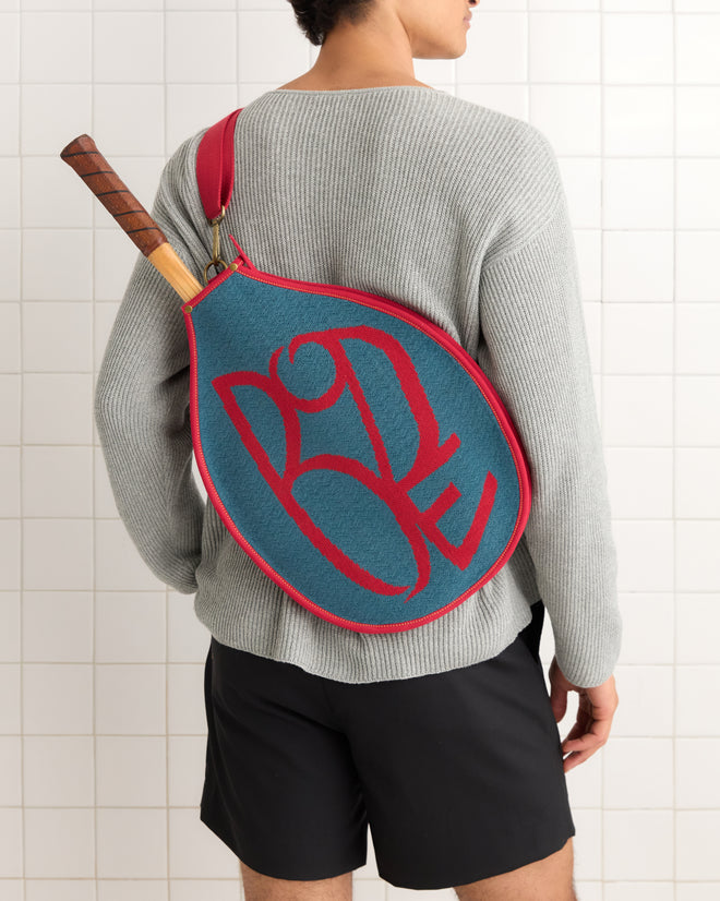 A person carries the Jacquard Racquet Sleeve over their shoulder, wearing a gray sweater and black shorts; the sleeve has an adjustable strap for comfort.