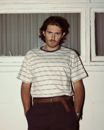 A person with shoulder-length hair and a mustache is standing against a white wall and window, wearing a Rec Jacquard Boxy Tee made of 100% cotton, dark pants, and a brown belt.