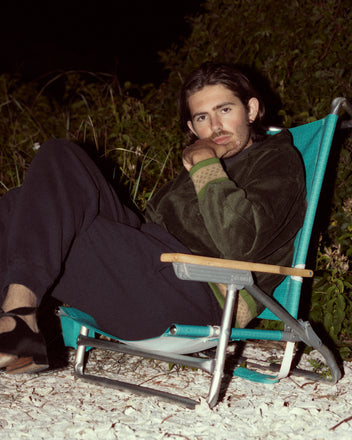 A person with long hair is sitting casually in a blue folding chair on a gravel area at night, wearing dark pants and the Peerie Rib Banbury Jacket.