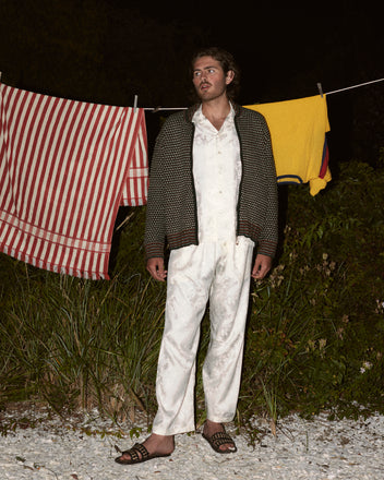 A man in pajamas stands outdoors at night, wearing a Schenectady Sweater over his pajamas. Behind him, a striped towel and a yellow towel hang on a clothesline.