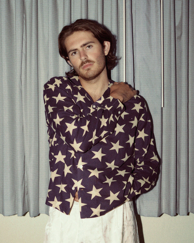 A person stands in front of blue curtains, wearing a 100% cotton Starry Long Sleeve Shirt adorned with intricate embroidered Bode details and paired with white pants, with one arm resting on their shoulder.