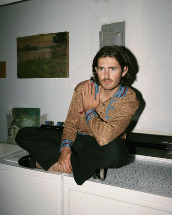 A person with long hair and a mustache, wearing the Goaltender Sweater in Gold and black pants, sits on a washing machine in a room with white walls adorned with a painting. A pair of hockey sticks are propped up against the wall, adding an unexpected touch to the scene.