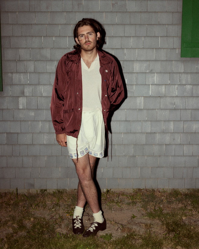 A person with long hair stands outside against a gray tiled wall, wearing a burgundy Nylon Track Jacket, white shirt, embroidered white shorts, socks, and brown shoes, with hands in pockets.