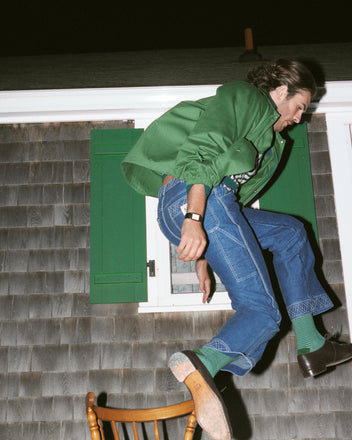 A person wearing blue jeans, a green Cotton Track Jacket, and green socks is jumping out of a window onto a wooden chair at night.