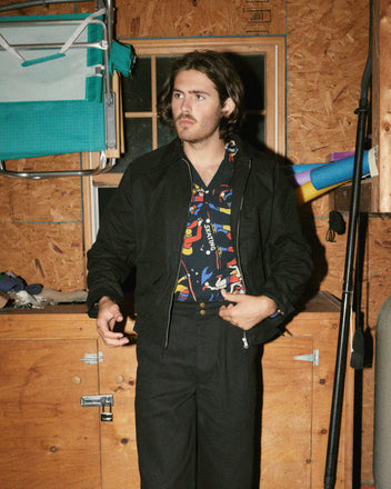 A man with long hair, wearing a patterned shirt and the Coach's Jacket in black, featuring front patch pockets, stands in a wooden workshop or garage with various tools and items hanging on the wall behind him.