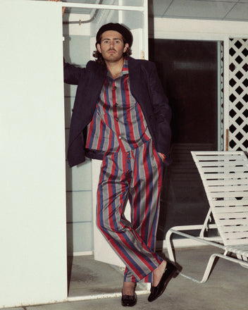 A person in Marina Stripe Pajama Pants and a blazer stands next to a door, leaning against the frame with one hand, in an outdoor setting with a lounge chair nearby. The classic pajama silhouette of their attire evokes both comfort and timeless style.