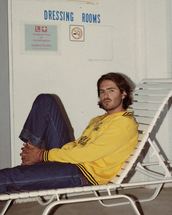 A person in a cozy, Club Long-Sleeved Polo - Yellow and blue jeans sits on a lounge chair in front of a wall with signs for dressing rooms and emergency information.