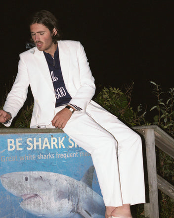 A man in a white suit sits on a sign, smoking a cigarette at night. The sign reads 