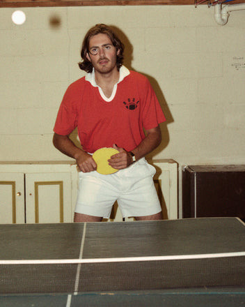 A man with long hair and a mustache plays table tennis indoors, sporting the vibrant red Worcester Polo paired with white shorts, holding a yellow paddle.