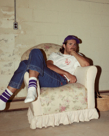 A person wearing an Embroidered Hoop Tee, blue jeans, a purple cap, and white socks with purple stripes featuring red embroidery, sits on a floral-patterned armchair in a room with unfinished walls. They are resting their head on their hand.