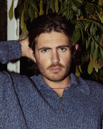 A person with long dark hair, a beard, and a serious expression, wearing the stylish Melange Polo with a subtle knit pattern and a ball-chain zipper, stands in front of leafy foliage with one hand behind their head.