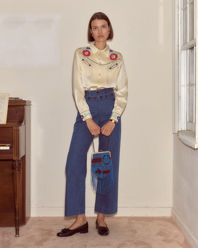 A woman in a cream western shirt and high-waisted jeans stands by a piano, holding the Capri Bag, a blue accessory adorned with seed beads.