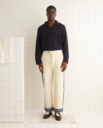 A person stands in a tiled room, wearing a dark sweater paired with the Caracalla Vine Trousers in undyed cream wool, alongside black shoes.