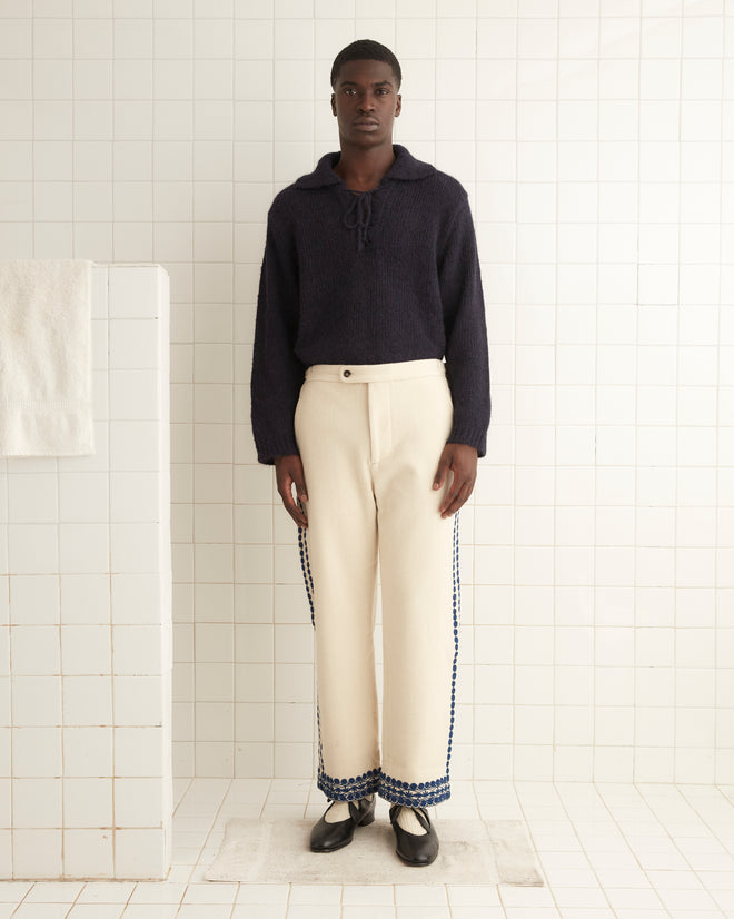 A person stands in a tiled room, wearing a dark sweater paired with the Caracalla Vine Trousers in undyed cream wool, alongside black shoes.