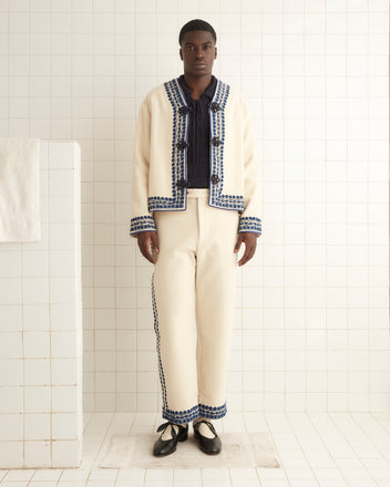 A man stands in a tiled room, wearing the Caracalla Vine Jacket, an embroidered piece in cream and blue, paired with black shoes, facing forward.