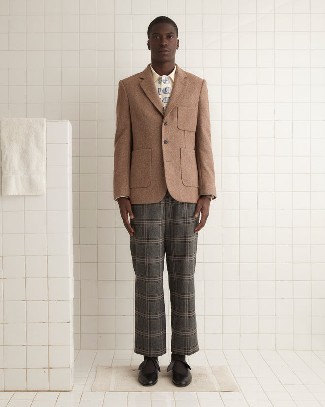 A person in a Carbon Print Suit Jacket - 40 and plaid pants stands confidently against a wooden wall, showcasing timeless style with suiting fabric that exudes elegance.