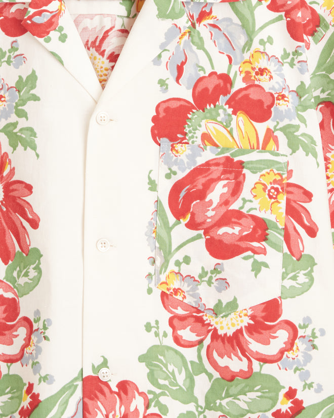 A Cardinal Flower Short Sleeve Shirt with a large, colorful floral pattern in red, green, blue, and yellow hangs on a wall hook, resembling a vibrant midcentury tablecloth.