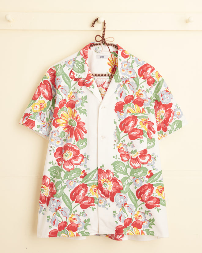 A Cardinal Flower Short Sleeve Shirt with a large, colorful floral pattern in red, green, blue, and yellow hangs on a wall hook, resembling a vibrant midcentury tablecloth.