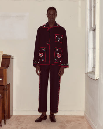 A person stands indoors wearing the Carroll Front Tab Jacket, an Alpine-style black piece with red trim and embroidered heart and flower appliqués.