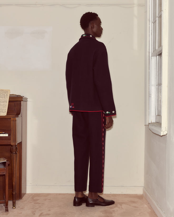 A man in a black suit with red trim wears the Carroll Front Tab Jacket, decorated with heart and flower appliqués, as he stands facing a cream wall near a window and an upright piano.