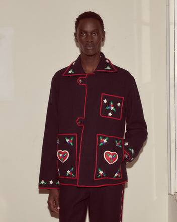 Wearing the Carroll Front Tab Jacket, featuring dark wool with red trim and heart and floral embroidery, a person stands against a plain wall, channeling whimsical Alice Through the Looking Glass style.