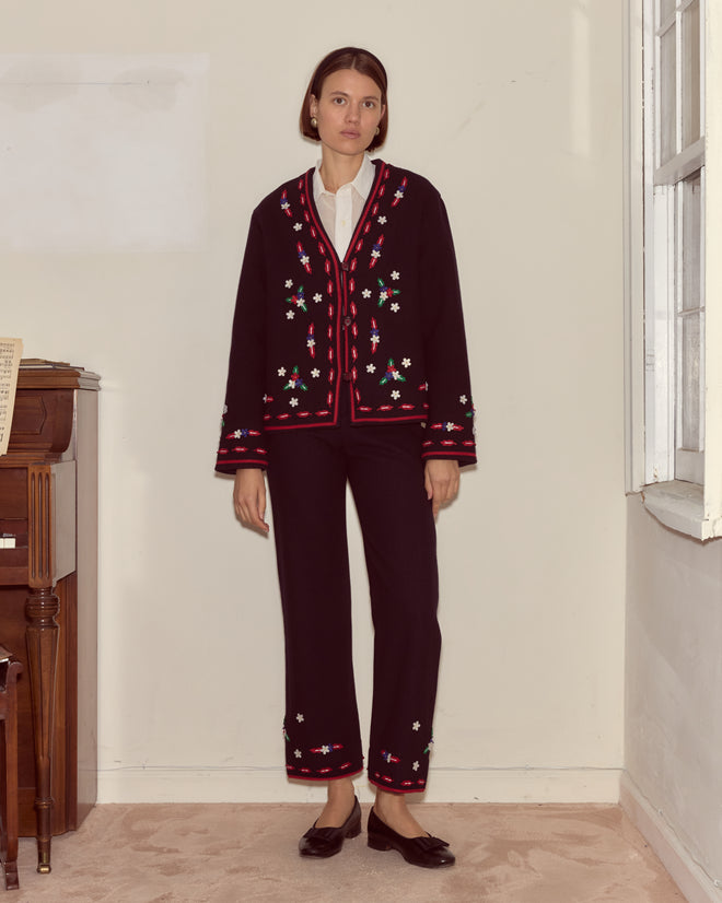 A woman wearing the Carroll Jacket, a black pantsuit with colorful flower appliqués, stands indoors near a piano and window.