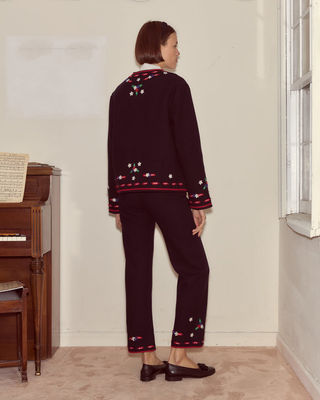 A woman stands indoors near a piano and window, facing away while wearing the Carroll Jacket—an elegant black embroidered suit adorned with floral appliqués.