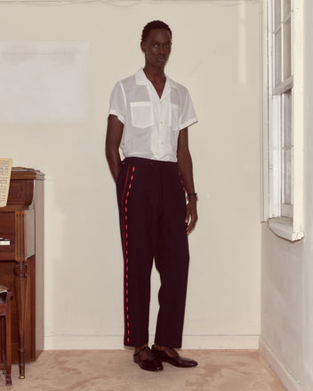 Wearing a white short-sleeve shirt and Carroll Side-Buckle Trousers, which are black wool trousers with red stripes, a person stands in a bright room.