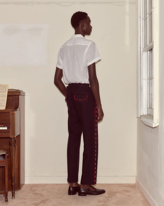 A person stands indoors facing a wall, wearing a white shirt and black Carroll Side-Buckle Trousers with red accents, evoking the style of Alice through the Looking Glass.