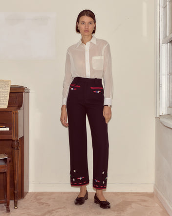 A woman in a white sheer blouse and Carroll Trousers with felted appliqués stands indoors by a piano and window.