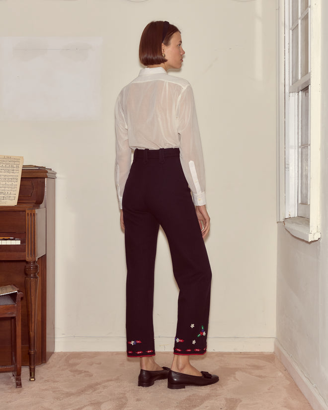 A woman wearing Carroll Trousers and a sheer white blouse stands facing away in a room with a piano and window.