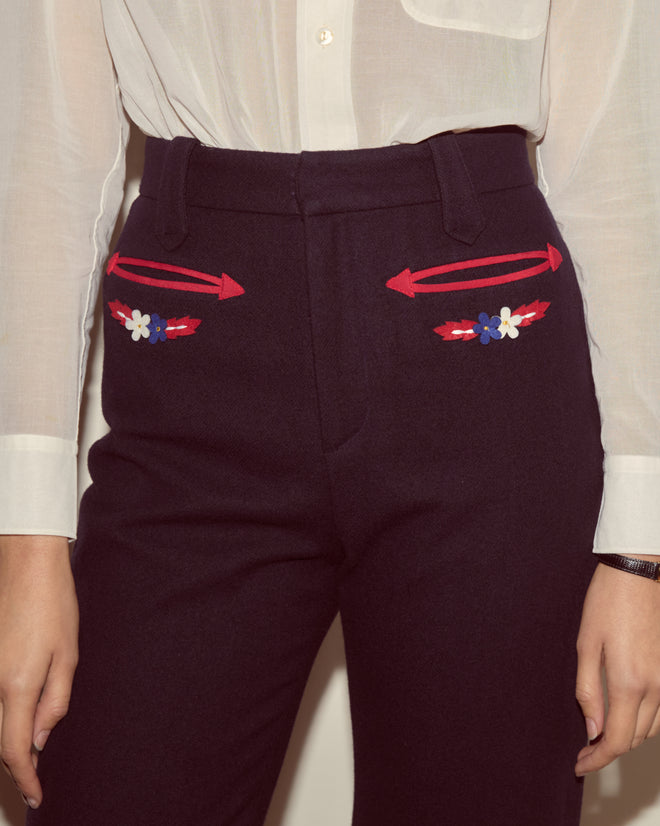 Wearing the Carroll Trousers—dark Western-cut pants with red arrows and small floral embroidery by the pockets—paired with a sheer white shirt for a subtle Alpine style.
