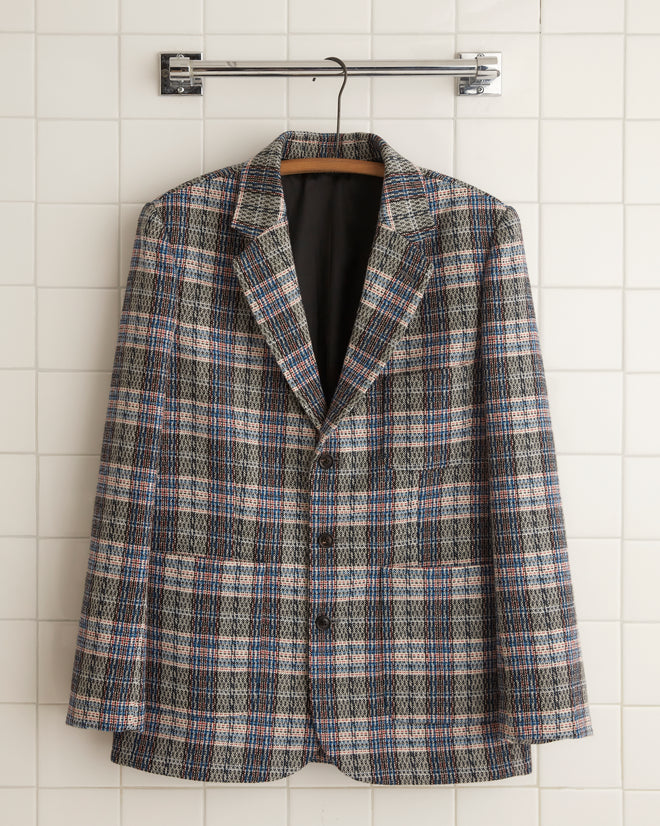 The Cartographer Plaid Suit Jacket - 44, made from luxurious 1960s-style suiting fabric, hangs elegantly on a hook against the white tiled wall.