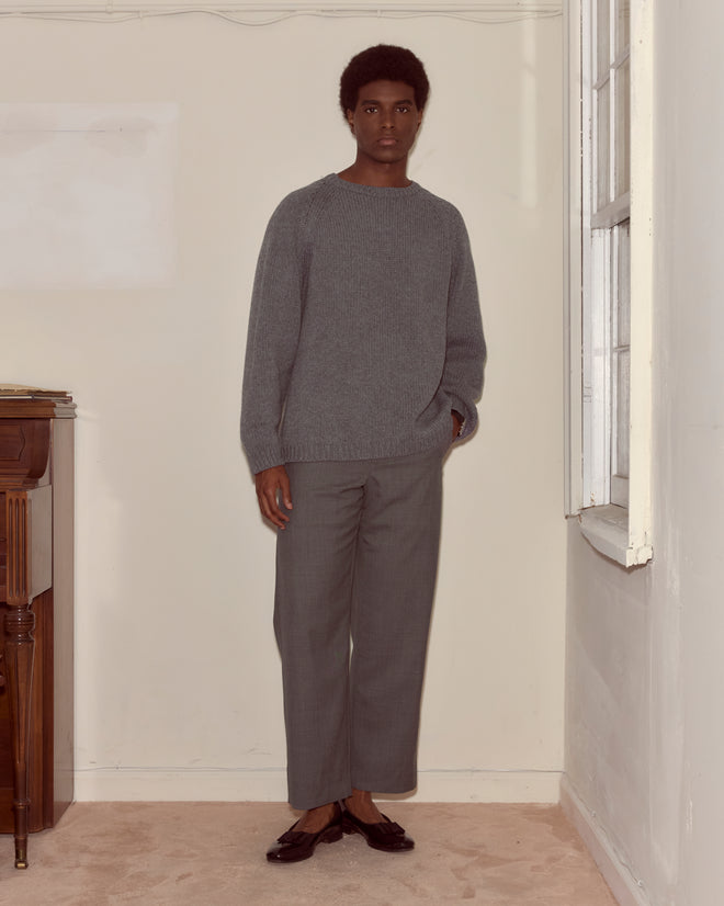 A person wearing the Cashmere Crewneck - Grey stands indoors by a window, looking at the camera.