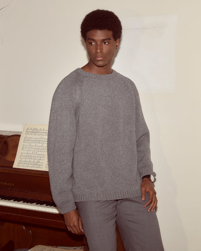 A man wearing a grey Cashmere Crewneck stands by a piano with sheet music, looking to the side.