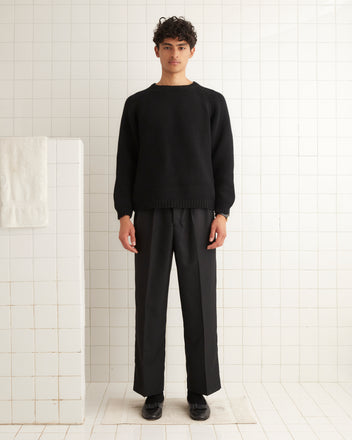 A person wearing a Cashmere Crewneck in black and pants stands in a tiled room next to a towel on a railing.