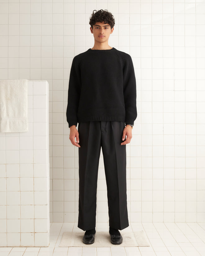 A person wearing a Cashmere Crewneck in black and pants stands in a tiled room next to a towel on a railing.