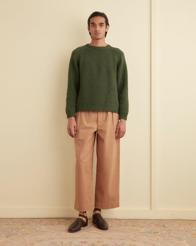 A person stands against a beige wall wearing an oversized green Cashmere Crewneck sweater, light brown trousers, colorful socks, and brown shoes.