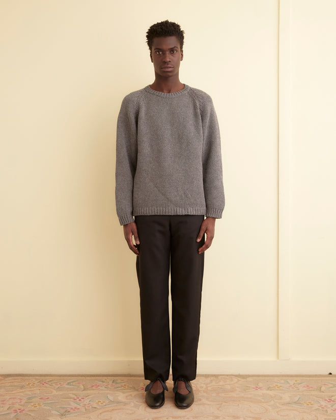 A person stands against a light cream wall, wearing the Cashmere Crewneck in grey with black pants and black shoes, looking directly at the camera.