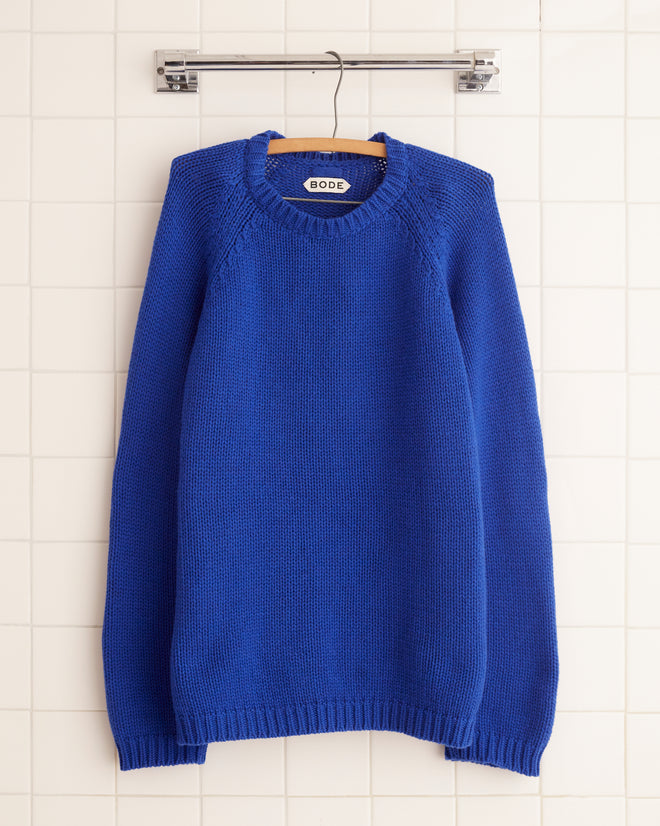 The Cashmere Crewneck - Lapis, featuring an oversized fit, hangs elegantly on a wooden hanger against a white tiled wall.