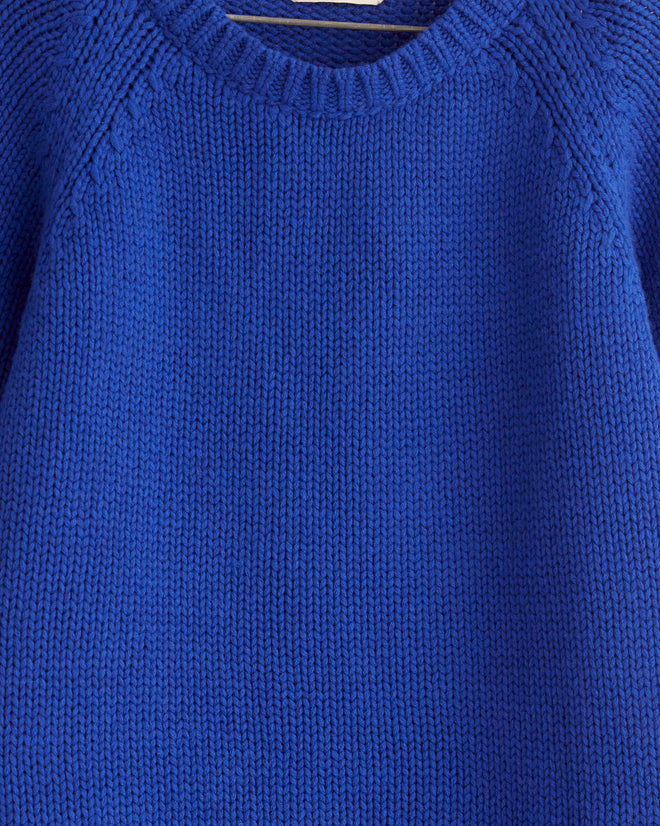 The Cashmere Crewneck - Lapis, featuring an oversized fit, hangs elegantly on a wooden hanger against a white tiled wall.