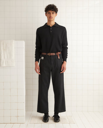 A young man wearing a black Cashmere Polo and a brown belt stands in front of a white tiled wall.