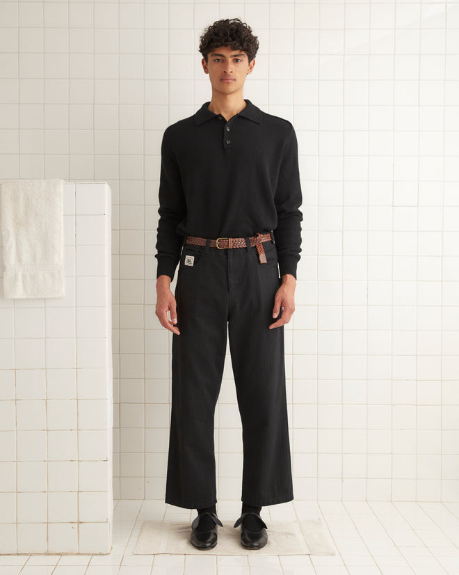 A young man wearing a black Cashmere Polo and a brown belt stands in front of a white tiled wall.