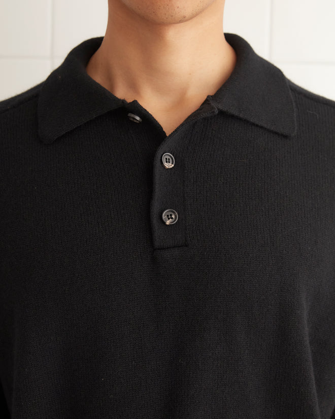 A young man wearing a black Cashmere Polo and a brown belt stands in front of a white tiled wall.